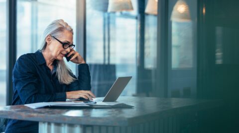 Woman gray hair glasses talking on phone looking at computer