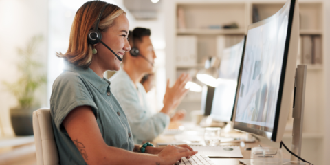 A female employee wearing a headset and short sleeve button up shirt smiles while looking at her computer screen.