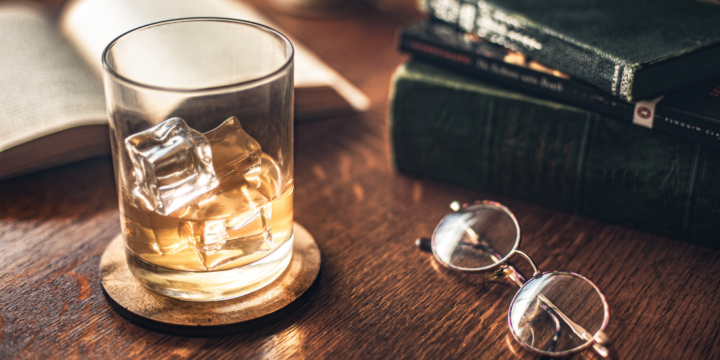 A glass of bourbon with ice on a coaster on a table with a pair of glasses, an open book and a stack of books near it.