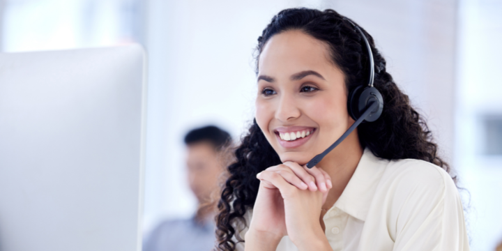 A contact center employee, with a headset on and hands clasped under her chin, smiles while listening and looking at her computer monitor.