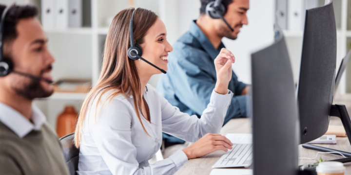 A contact center agent sits in a row of agents, smiling while talking on her phone headset.