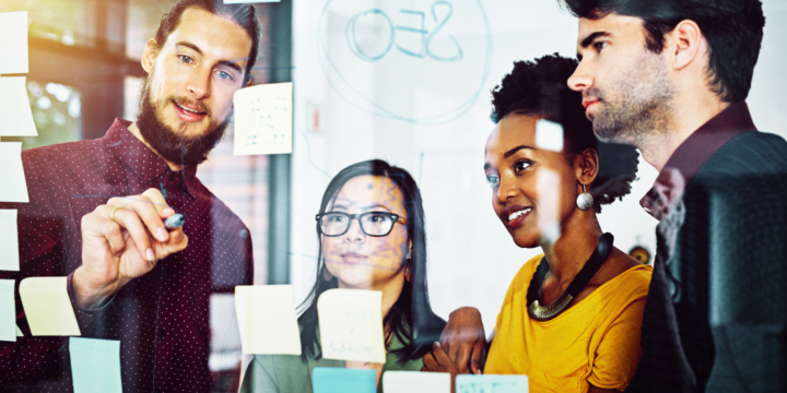 Two men and two women collaborate on sticky notes on a glass wall in the office.