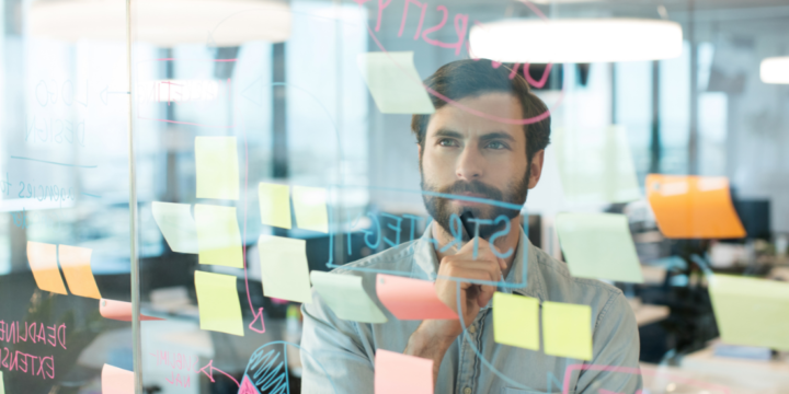 A white man with a beard ponders charts and sticky notes in an office setting.