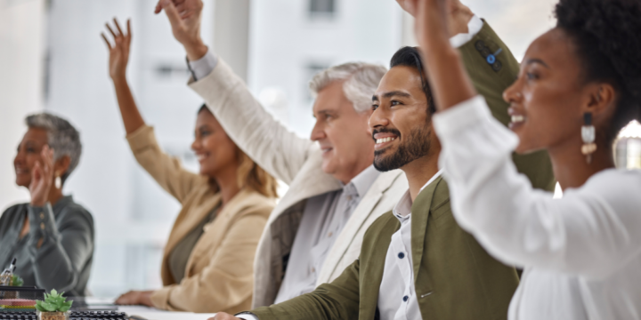 A diverse group of happy looking employees sit at a table, all with hands raised to engage with the meeting they're in.