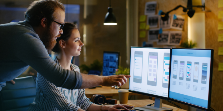 A woman and man look at a set of two computer screens together. On the screen are mock ups of an mobile app user experience design.