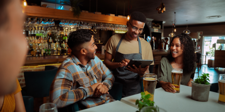 A waiter holding a tablet serves a pair of smiling customers in a restaurant.