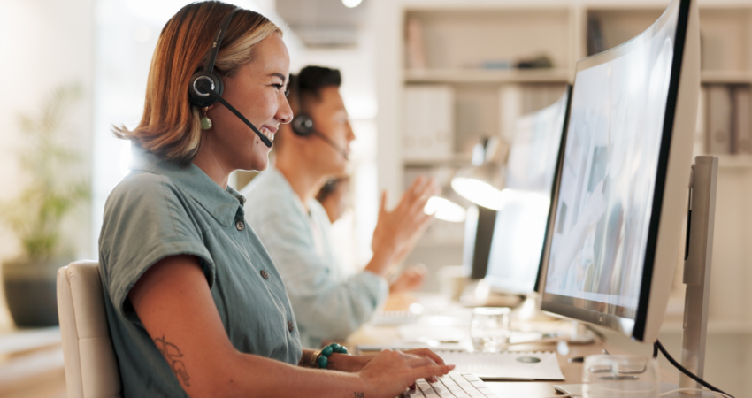 A female employee wearing a headset and short sleeve button up shirt smiles while looking at her computer screen.